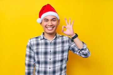 Fototapeta na wymiar Smiling young Asian man wearing a plaid shirt in a Christmas hat posing showing okay ok gesture on yellow background. celebration Christmas holiday and New Year concept