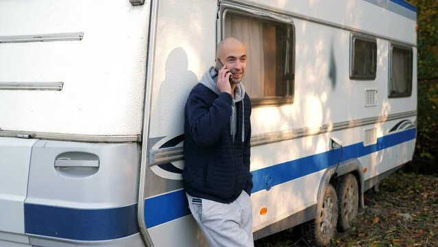 A man on vacation in the autumn forest is talking on the phone standing near a mobile van, camping with a motorhome in the autumn forest. Active leisure.