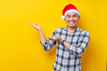 Cheerful young Asian man in Santa hat showing hand aside at blank space for advertising text on yellow studio background. celebration Christmas holiday and New Year concept