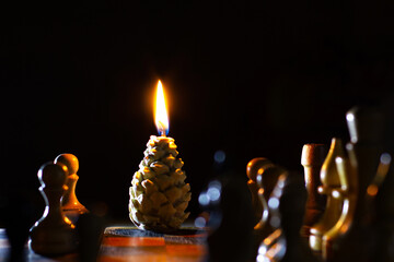 Burning candle in pine cone shape. Chess in the foreground. Handmade. Holiday advent. A candle on a...