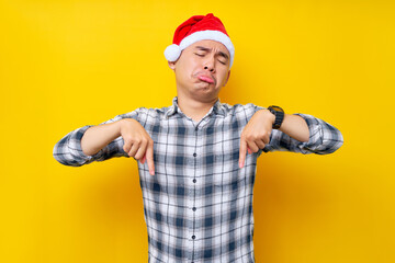 Annoyed young Asian man wearing a plaid shirt in a Christmas hat pointing finger down indicate the workspace area on yellow background. celebration Christmas holiday and New Year concept