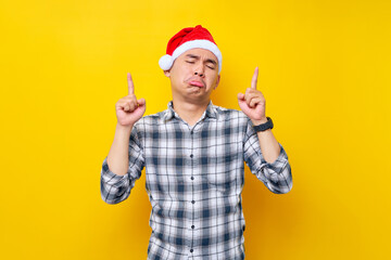 Fototapeta na wymiar Sad young Asian man wearing a plaid shirt in a Christmas hat pointing finger overhead on the workspace area on yellow background. celebration Christmas holiday and New Year concept