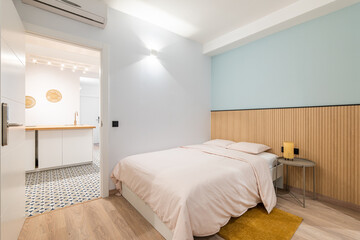 Bright room with a double bed, a bedside table and a terry yellow rug. Simple and minimalistic bedroom interior design in light pastel colors. The doorway reveals a kitchen with modular furniture.