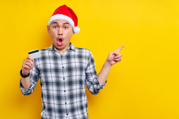 Fototapeta na wymiar Surprised young Asian man wearing Christmas hat holding credit bank card pointing index finger aside on copy space area on yellow background. celebration Christmas holiday and New Year concept