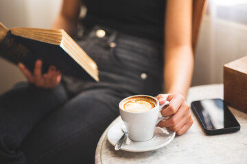 Young woman reading a book and holding a cup of coffee at home by the window