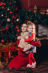 A young mother hugs her daughter and a teddy bear against the background of a Christmas tree