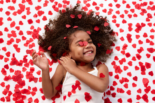 Funny young girl with afro hair laughing on bed surrounded by heart shaped confetti
