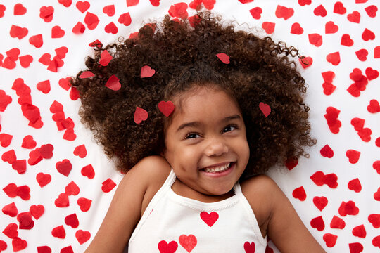 Happy young girl with afro hair giggling on bed surrounded by heart shaped confetti