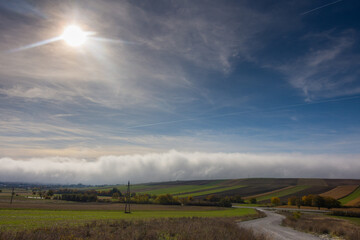 shining sun on blue sky over a white wall of dense fog on the ground with fields
