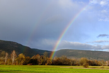 rainbow on dark rain clouds over green fields with trees and mountains