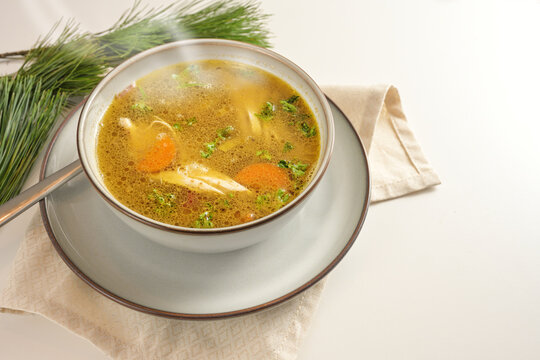 Hot steaming winter soup from chicken, vegetables and parsley in a bowl, home remedy against cold and flu, served on a gay white table with pine branches, copy space, selected focus