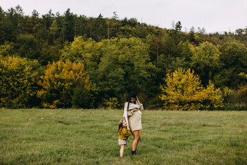 Pregnant woman and her toddler daughter outdoors, walking in a field with green grass. Little girl hugging her mother, spending time together.