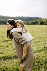 Mother giving a piggyback riding to her toddler daughter, outdoors, in a field. Mother and daughter spending time together, having fun.