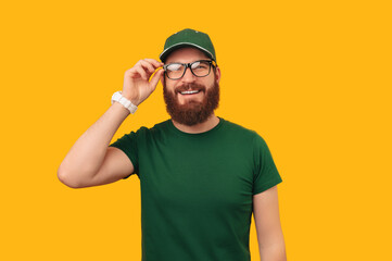 Fototapeta premium Portrait of a smiling delivery man wearing green uniform puts on some glasses over yellow background.