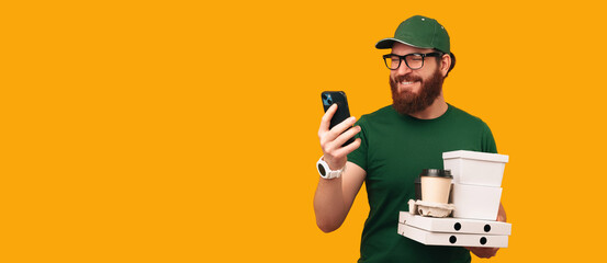 Delivery man watches the order on the phone and holds lunch boxes and cups. Studio shot with copy space.