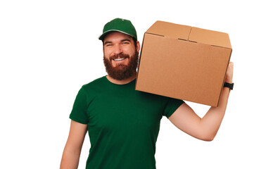 Strong delivery man wearing green uniform holds a carton box over his shoulder over white...