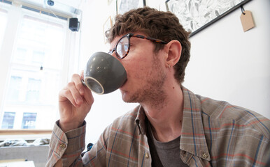 A young student hipster wearing glasses, drinking a coffee cappuccino in a trendy coffee bar in the city. Student preparing for the days long lectures. Coffee stimulant for the day ahead.