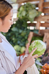 Obraz premium Grocery store, lettuce and Asian woman shopping and looking at vegetable quality and sale. Customer holding and checking vegetables price on salad promotion in a health food shop or supermarket