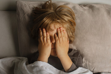 The child was scared before going to bed. Night terrors in a child. The kid covers his face with his hands in a fear. Children's experiences. Boy in the bed. View from above. Sad psychological state.