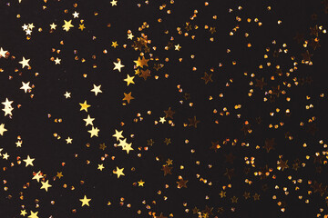 Glittering gold colored stars and crystals confetti on a black background. Festive composition. Selective focus.