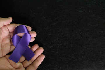 Human hand holding purple or violet ribbon color on dark black background. National cancer prevention month, testicular and pancreatic cancer awareness and hope concept.