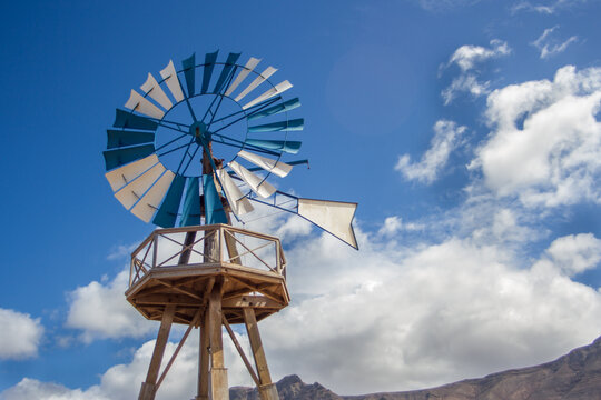 Desertic landscape. Windmill with blue and white blades. Mountains in the background. Yellow sand dunes with desert plants. Blue sky with white fluffy clouds. Famara Beach, Lanzarote, Canary Islands, 