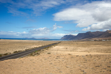 Desertic landscape. Lone black road runs through a desert plain. Mountains in the background. Blue sky with large white fluffy clouds. Yellow sand dunes with desert plants. Famara Beach, Lanzarote