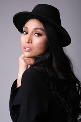 Fashionable brunette asian ethnicity in stylish black coat and hat od grey background.