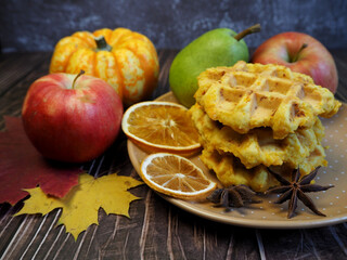three pumpkin Belgian waffles without gluten from corn flour lie on a gray plate with three slices of dry orange and two stars of anise against the background of pumpkin apples and pears on a wooden t