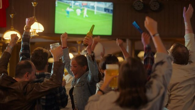 Group of Soccer Fans Cheering, Screaming, Raising Hands and Jumping During a Football Game Live Broadcast in a Sports Pub. Player in Blue Shirt Scores a Goal and Friends Celebrate. Slow Motion Footage