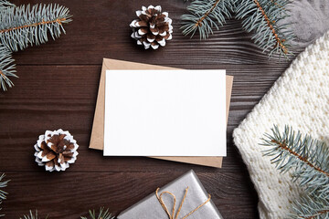 Christmas greeting card mockup with envelope, gift box, green fir tree branches and holiday decorations on brown wooden background, top view, flat lay. White New Year card with decor