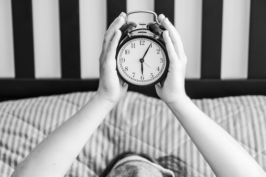 Person lying in bed holding an alarm clock in the hands at six o'clock in the morning, laziness concept, black and white image