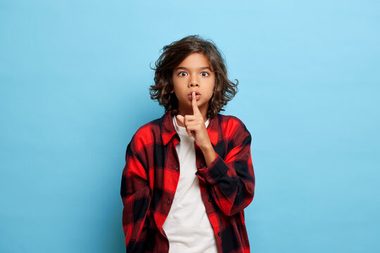 Indoor shot of amazed young teen boy with curly hair shows silence sign, keeps finger on lips, wears casual red shirt, poses over blue wall