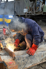 A worker welding metal parts on a construction site. A welder welds parts of a large machine in an open pit coal mine. An interesting example of manual work.