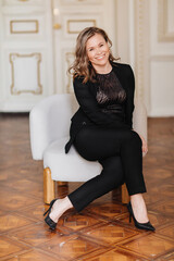 an attractive woman in a black pantsuit and heels in a chair in classic interior