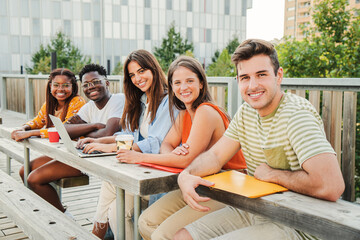 Multiracial group of happy teenage students looking at camera, smiling relaxed together outside the university campus taking a break. Meeting of young and fun college people enjoying the sunlight