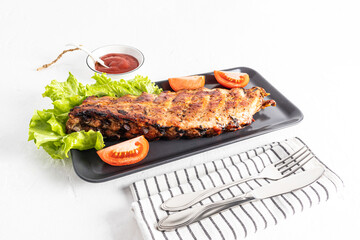 Grilled pork ribs are served on a rectangular dark ceramic plate with fresh vegetables and a bowl of sauce. white background. recipe menu.