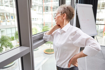 Naklejka na ściany i meble Elderly businesswoman stands thoughtfully by the window