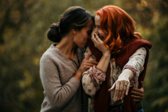 Two Beautiful Lesbian/gay Females Announce To Their Friends That They Are Engaged. Redhead Woman Showing An Engagement Ring, While Her Partner Is Hugging Her And Smiling.