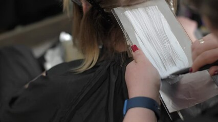 Master's hand applies hair dye with brush on strand of female hair on special sheet of foil, copy space. Process of highlighting by professional master in beauty salon. Paint hair close-up