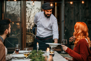 A male chef in uniform is serving a private elegant dinner to a group of people, People are smiling and welcoming the chef who is serving and bringing delicious food.