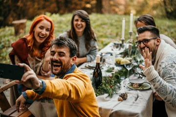 A Group of people having a garden party dinner, and a man is holding a smartphone and taking a group selfie with his friends.
