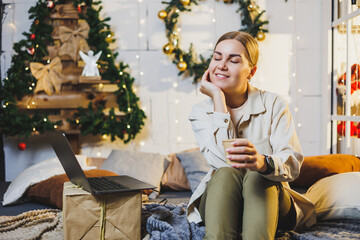 Pretty woman with blond hair in cozy clothes with coffee in hand using laptop working remotely and smiling on bed in room with Christmas tree at home. New Year holidays.