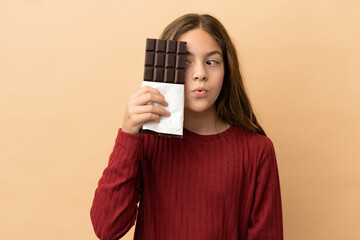Little caucasian girl isolated on beige background taking a chocolate tablet and surprised