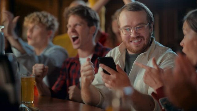 Portrait of a Happy Young Man Holding a Smartphone, Anxious About the Sports Bet on a Soccer Match. Looking at the Screen While Sitting at a Counter, Getting Very Emotional After Winning the Bet.
