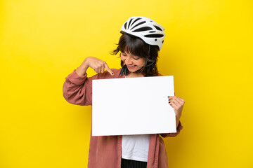 Young cyclist latin woman isolated on yellow background holding an empty placard with happy expression and pointing it