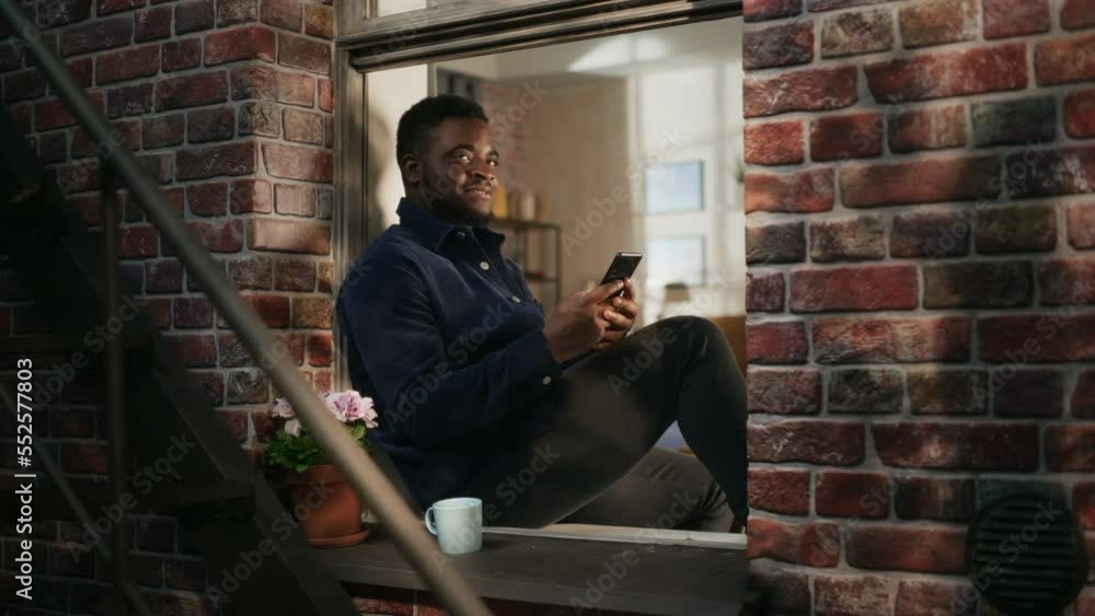 Aufkleber black authentic man using smartphone in his flat while sitting on bedroom windowsill. african americ