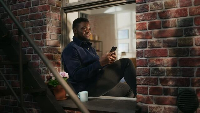 Black Authentic Man Using Smartphone in his Flat While Sitting on Bedroom Windowsill. African American Male Posting Photos on Social Media and Using Filters While Smiling. Calm and Relaxing Evening.