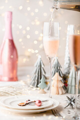 Xmas table settings in white and pink colors with trendy paper christmas trees on background. Table ready for festive dinner and party with pink prosecco pouring to the glass