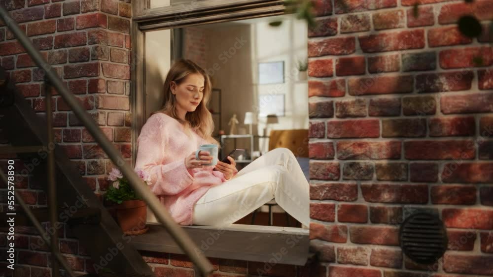 Poster woman in pink comfy sweater using smartphone and drinking herbal tea while sitting on her windowsill